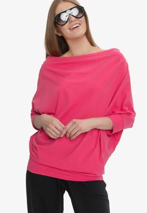 Kobieta w oversize jasnoróżowym topie, czarnych okularach przeciwsłonecznych i czarnych spodniach, uśmiechająca się z rękami splecionymi z przodu.