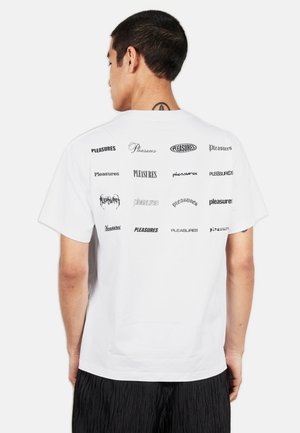 Mężczyzna z krótkimi ciemnymi włosami, ubrany w białą koszulkę z wieloma logo "Pleasures" w różnych czcionkach nadrukowanych na plecach.