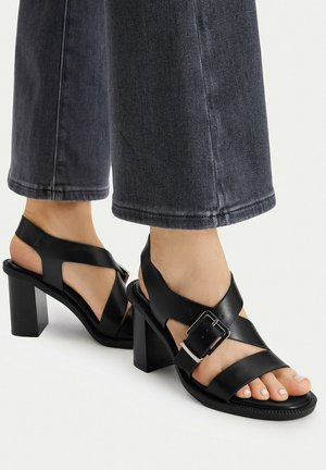 Schwarze Leder-Sandalen mit Blockabsatz, breiten Kreuzriemen und großer silberner Schnalle, getragen zu dunklen, gekürzten Jeans.