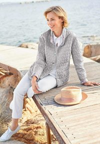 Grau gesteppten Jacke mit einem blumenmusternden Kragen, getragen über einem weißen Hemd und Hose. Ein Strohhut mit einem pinken Band liegt auf dem Holzsteg.