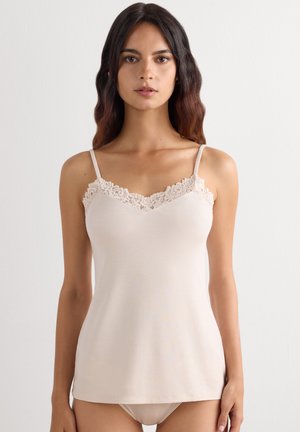 Vrouw draagt een beige hemdje met kanten randje en bijpassend ondergoed, staand tegen een effen lichte achtergrond.