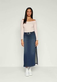 Top off-the-shoulder em rosa claro combinado com uma saia maxi de denim azul escuro com fenda lateral, detalhe de cinto e ténis brancos.