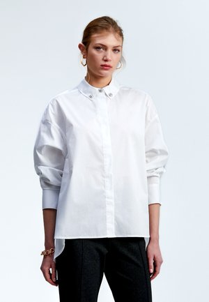White long-sleeve shirt made of cotton blend, featuring a high collar and decorative buttons, with a relaxed fit and cuffed sleeves.