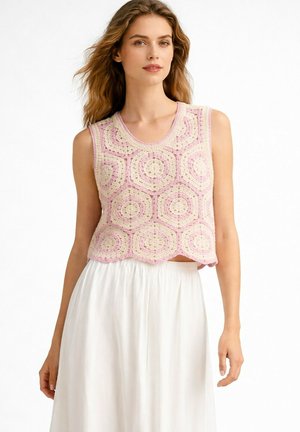 Femme portant un haut crocheté sans manches rose et crème avec des motifs hexagonaux, associé à une jupe blanche fluide, debout devant un fond blanc.