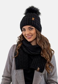 Bonnet noir en tricot à torsades avec un pompon en fausse fourrure, assorti à une écharpe noire. Les deux accessoires présentent un motif texturé et une étiquette en cuir.