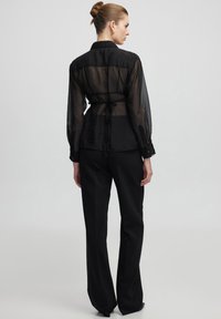 Blusa nera trasparente con maniche lunghe, polsini a bottone e un dettaglio con fiocco sul retro. Indossata con pantaloni neri a vita alta.