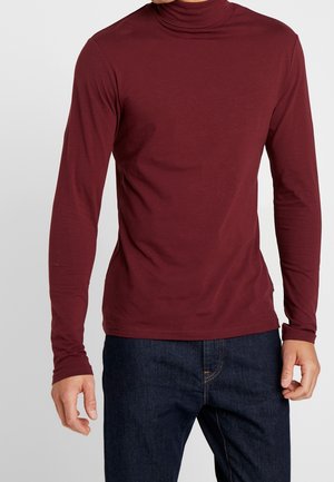Homme portant un col roulé ajusté à manches longues bordeaux avec un jean bleu foncé, visible du cou aux hanches.
