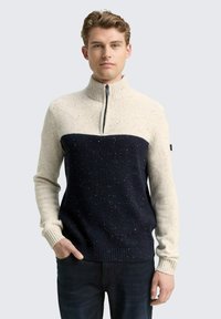 Junger Mann in einem Halb-Reißverschluss, zweifarbigen Sweater in Creme und Marineblau mit gesprenkelter Textur, der mit einer Hand in der Tasche vor einem schlichten Hintergrund steht.