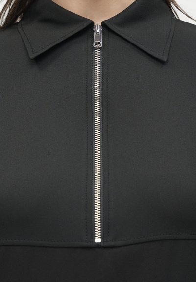 Κοντινό πλάνο ενός μαύρου ενδύματος με γιακά και ασημένιο φερμουάρ στο κέντρο, εν μέρει φερμουαρμένο.