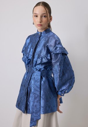 Fiatal nő, aki fényes kék, texturált, öves dzsekit visel fodros részletekkel és dús ujjakkal, egy egyszínű szürke háttér előtt pózol.