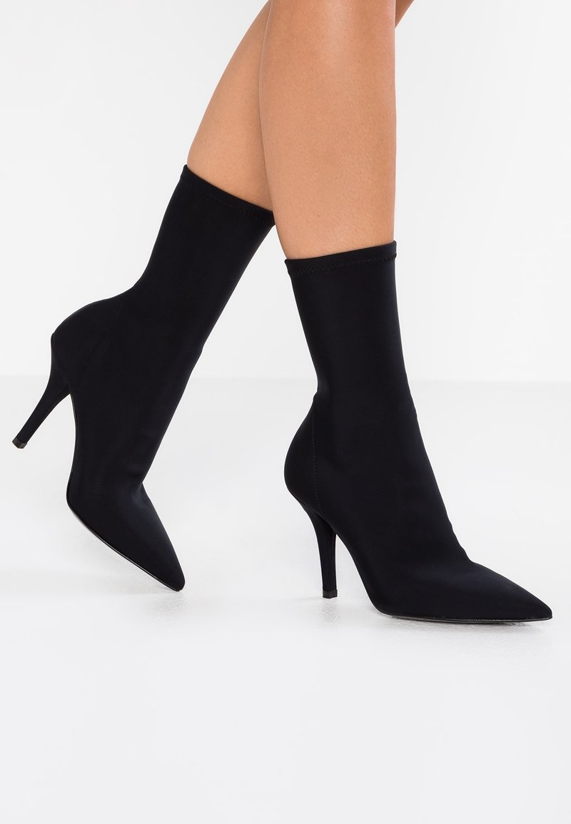 Jambes portant des bottines à bout pointu noires en forme de chaussette avec des talons hauts fins, présentées sur un fond blanc.