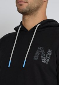 Musta huppari, jossa on kuvioitu "N"-logo, harmaat narut sinisillä päillä ja teksturoitu kangas. Läheinen kuva kaula-aukosta.