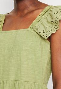 Lichtgroene jurk met een vierkante halslijn en gerimpelde, geborduurde mouwen. De stof heeft een subtiele structuur en een vloeiende silhouet.