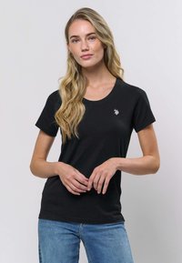 Črna bombažna majica s klasičnim okroglim izrezom, kratkimi rokavi in majhnim vezenim emblemom na levi strani prsnega koša.