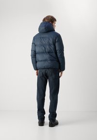 Marineblauwe pufferjack met een hoge kraag en capuchon, met doorgestikte delen. Draagt met donkere jeans en zwarte schoenen.