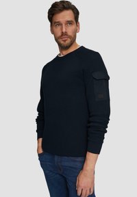 Navyblauwe gebreide trui met ribbeltextuur, ronde hals en een zijzakdetail op de linkermouw. Gecombineerd met denim jeans.