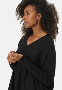 Zwarte v-hals trui met lange mouwen, zachte textuur en een relaxed fit. Krullend haar is zichtbaar op de schouders en handen van het model.