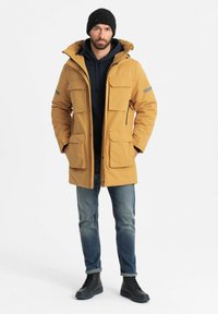Geel geïsoleerde parka met meerdere buitenzakken, ritssluitingen en een afneembare capuchon, gedragen over een marineblauwe hoodie, gecombineerd met denim en zwarte laarzen.