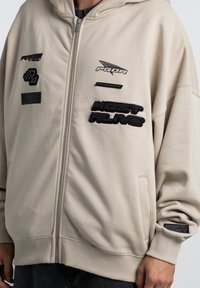 Beige zip-up hættetrøje med forskellige sorte lapper, strukturerede detaljer og sidelommer. Fremstillet af blødt stof med en afslappet pasform.