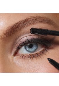 Acercamiento de un ojo azul con pestañas largas y oscuras. Un cepillo de máscara negra está aplicando el producto. Se pueden ver sombras de ojos rosa suave y una textura de piel suave.