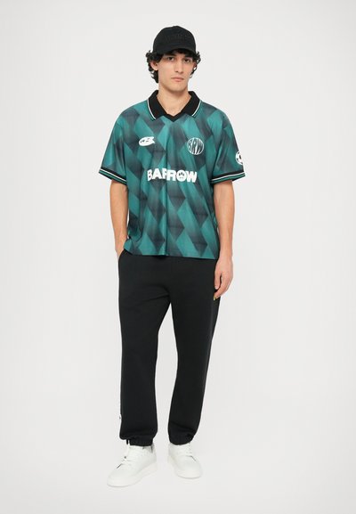 Polo shirt à motifs verts et noirs avec un col, porté avec un pantalon de jogging noir et des baskets blanches. La casquette présente une inscription en relief.