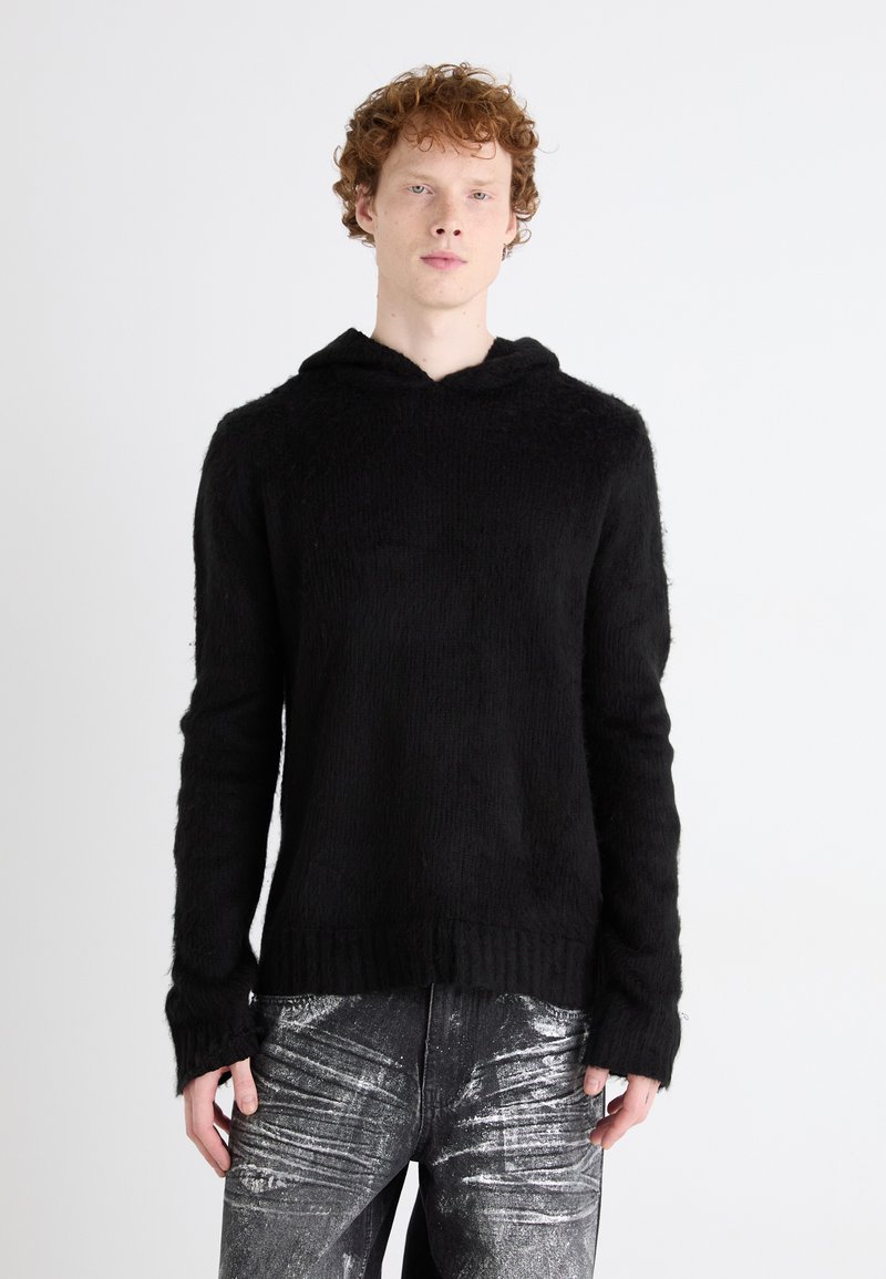 Hoodie preto de malha com um acabamento suave e texturizado. Apresenta uma gola larga e mangas longas com punhos canelados. Combinado com jeans com detalhes em prata.