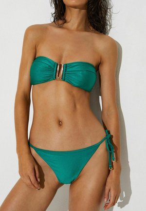 Kvinde iført en grøn strapless bikini med en metalspænde detalje på toppen og side-bindebund på en hvid baggrund.