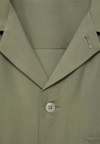 Blazer verde măsliniu cu rever în formă de breton, închidere cu un singur buton și un buzunar pe pieptul stâng. Materialul are un finisaj neted, texturat.