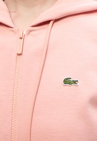 Κοντινό πλάνο ατόμου που φοράει ροζ φούτερ με φερμουάρ, με μικρό πράσινο λογότυπο κροκοδείλου και ταίριαστο τράβηγμα φερμουάρ.