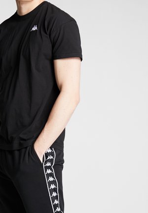 Svart T-shirt med kort ärm, tillverkad av bomull, med en liten logotyp på bröstet, parat med svarta byxor som har ett mönster av sidologotyper.