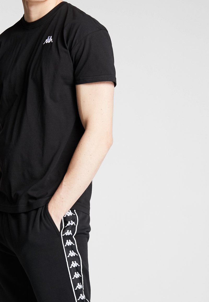 Černé triko s krátkým rukávem vyrobené z bavlny, které má malou nášivku na hrudi, a je v kombinaci s černými kalhotami se vzorem loga po straně.