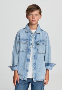 Jasnoniebieska kurtka dżinsowa z dwoma kieszeniami na piersi, zapinana na guziki, z podwiniętymi rękawami, noszona na białej koszulce z krótkim rękawem i zestawiona z niebieskimi dżinsami.