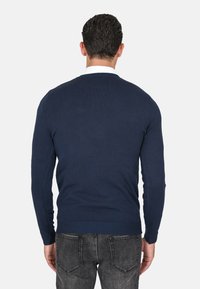 Maglione in maglia blu navy con scollatura rotonda e maniche lunghe, caratterizzato da un orlo e polsini a coste. Indossato con jeans grigio scuro, vista da dietro.