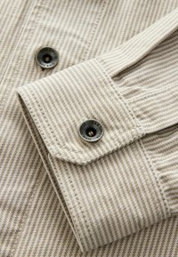 Gros plan sur le poignet d’une chemise rayée beige et blanche avec des boutons noirs étiquetés "Denim Supply Co" et une couture détaillée.