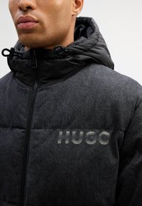 Svart hettejakke med puffersnitt og en teksturert grå kropp, med glidelås, og "HUGO"-logo i en lysere nyanse på brystet.