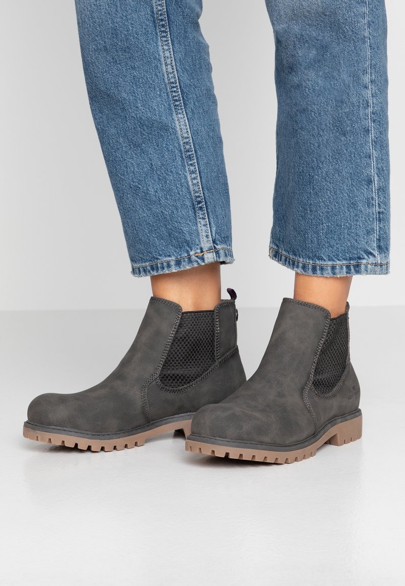 Osoba oblečená v šedých kotníkových botách s černými elastickými stranami a béžovými podrážkami se vzorkem, v kombinaci s zkrácenými modrými džínami.