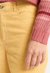 Gula kordbyxorna med vertikala ribbor, åtföljda av en texturerad rosa stickad tröja. En hand med en tatuering vilar på byxorna.