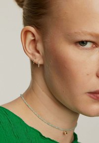 Pendientes de aro de oro con pequeños diamantes, un collar corto de cuentas en azul claro y una blusa verde texturizada con escote en V.