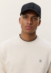 Casquette noire avec logo "II" en relief, portée par une personne en sweat-shirt beige clair à col rond et au design minimaliste.