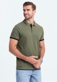Olivově zelené polo tričko s černým límcem a akcenty na rukávech, tříbuttonová légou, krátké rukávy a dekorativní prošívání na lemu.
