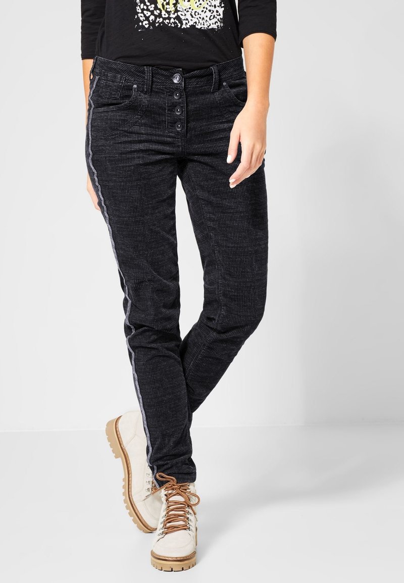 Femme portant un pantalon noir texturé avec bande latérale et des bottes blanches à lacets sur fond uni.