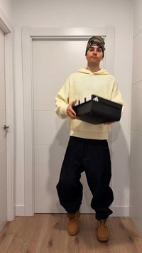 Osoba u kamuflažnoj kapi, kremastom džemperu i tamnim širokim hlačama drži crnu kutiju za cipele u bijelom unutarnjem hodniku.