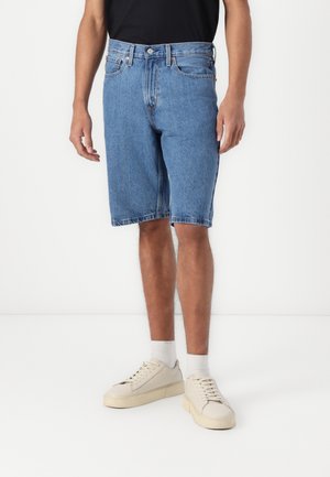 Homme portant un short en jean bleu mi-long, des chaussettes blanches, des baskets écru et une chemise noire, debout devant un fond blanc.