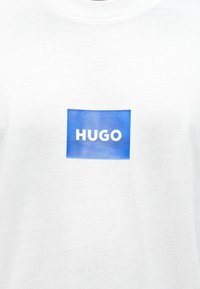 Biele bavlnené tričko s obdĺžnikovým modrým logom, na ktorom je výrazné biele slovo "HUGO" umiestnené v strede na hrudi.