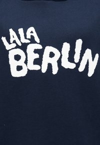 Tmavomodré bavlnené tričko s výrazným bielym nápisom. Nápis na prednej strane znie „LALA BERLIN“ v hravom, ručne kreslenom štýle.