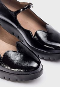 Chaussures noires en cuir verni avec un design festonné, bout rond et fine bride de cheville. Elles présentent une semelle épaisse en caoutchouc noir avec un relief texturé.