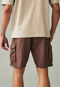 Braune Cargo-Shorts mit seitlichen Taschen, aus einem strukturierten Stoff gefertigt. Getragen mit einem hellbeigen Hemd mit kurzen Ärmeln.