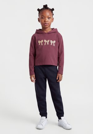 Jeune fille avec des cheveux tressés portant un sweat à capuche bordeaux avec trois nœuds à motif léopard, pantalon noir et baskets blanches, debout face à l'avant.
