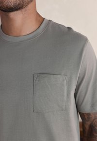 Graues Kurzarm-T-Shirt aus weichem Stoff, mit einer Brusttasche und präziser Naht. Runder Ausschnitt und lockere Passform.