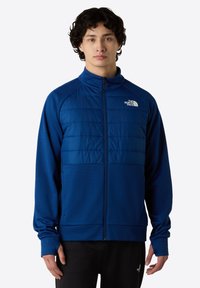 Modrá zipová bunda s polstrovanou přední částí, texturovanými rukávy, stojáčkem a logem na levé hrudi.